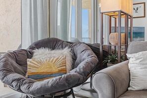 A modern living room with a grey upholstered chair, a small table with a woven basket, and a view of the ocean through large windows. - Oceania Plaza 502 (New Smyrna Beach)