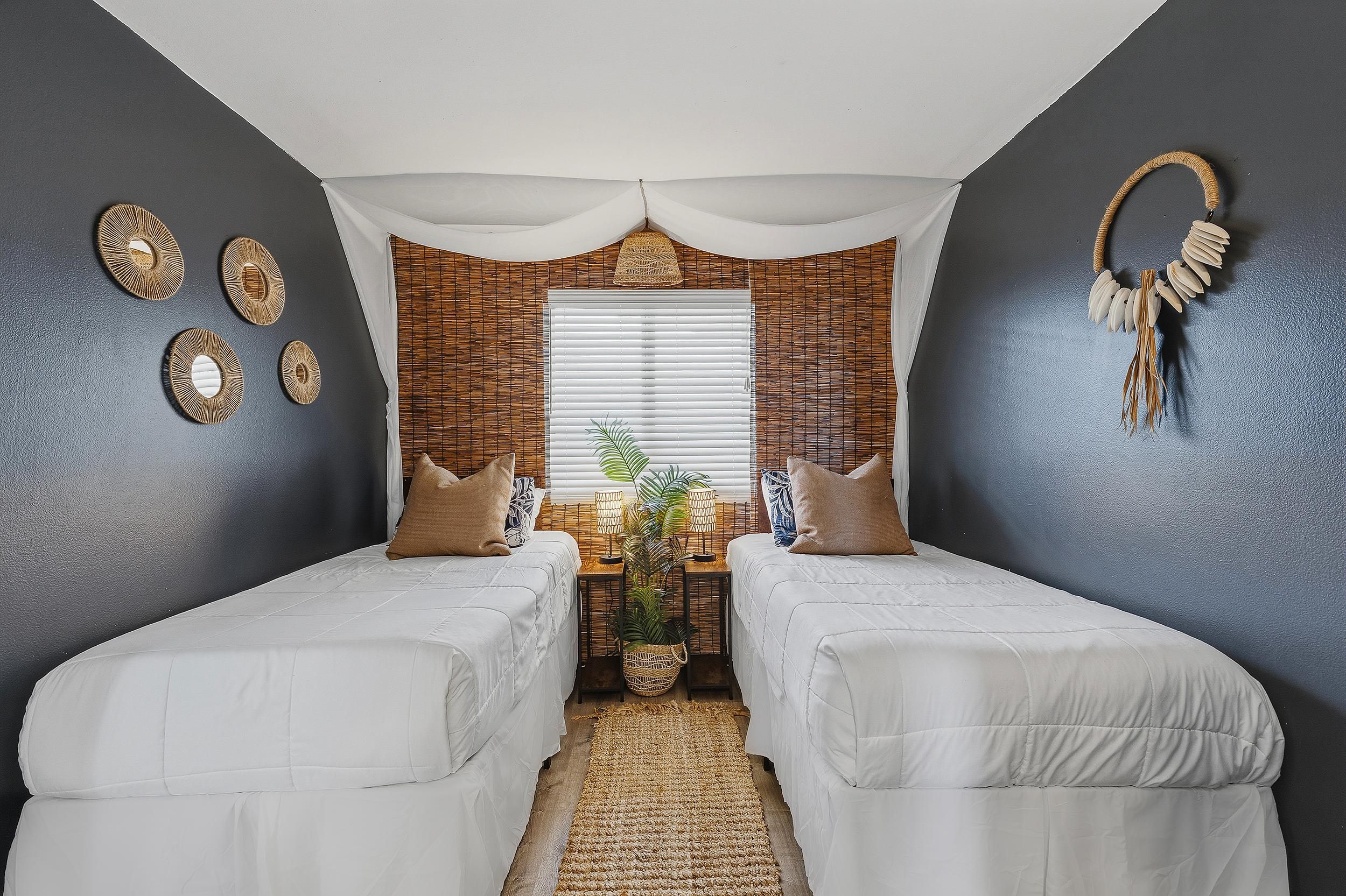 A room with two beds, a window with blinds, a woven headboard, and wall decor.