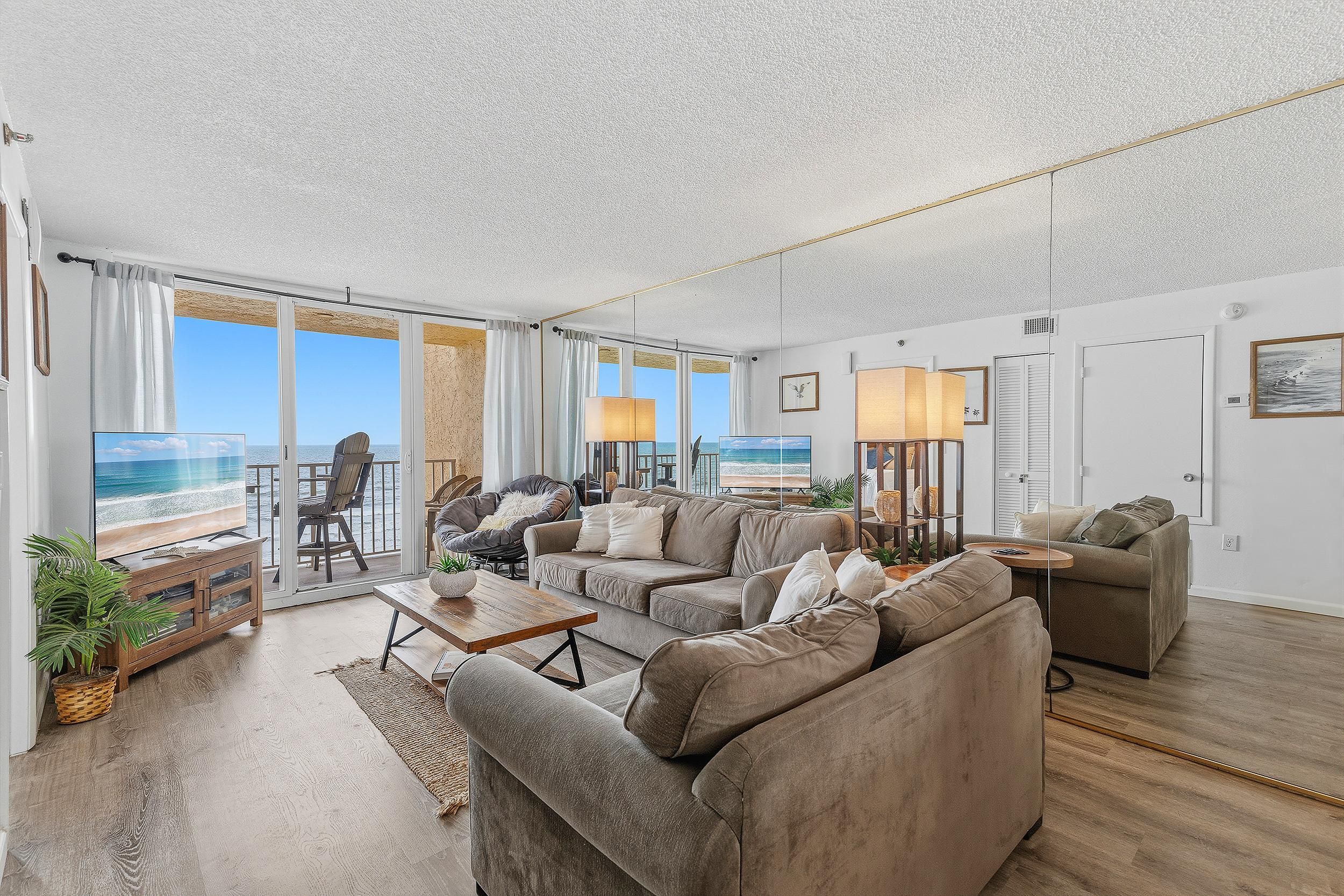 A spacious living room with a large window overlooking a beach, furnished with sofas and a coffee table.