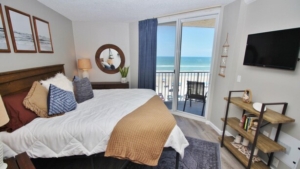 A bedroom with a bed, a bedside table, a lamp, a mirror, a TV, a balcony with a view of the beach, and a bookshelf. - Oceania Plaza 502 (New Smyrna Beach)