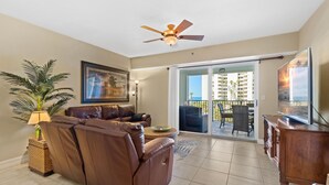 A living room with a brown leather sofa, a ceiling fan, a television, and a view of a balcony with outdoor furniture. - Oceanwalk 13-406 (New Smyrna Beach)