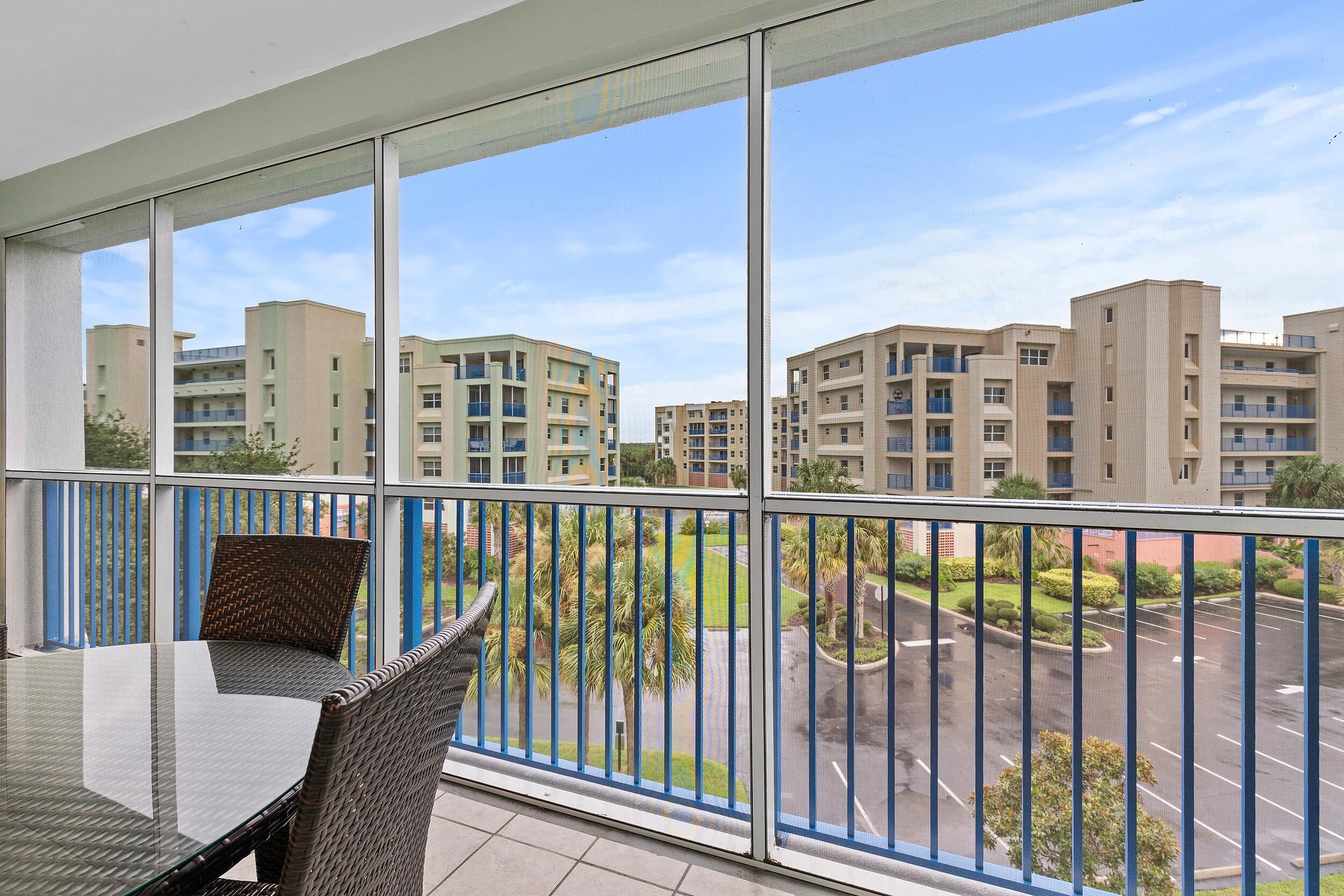 A balcony with a glass table and wicker chair, overlooking a parking lot and residential buildings.