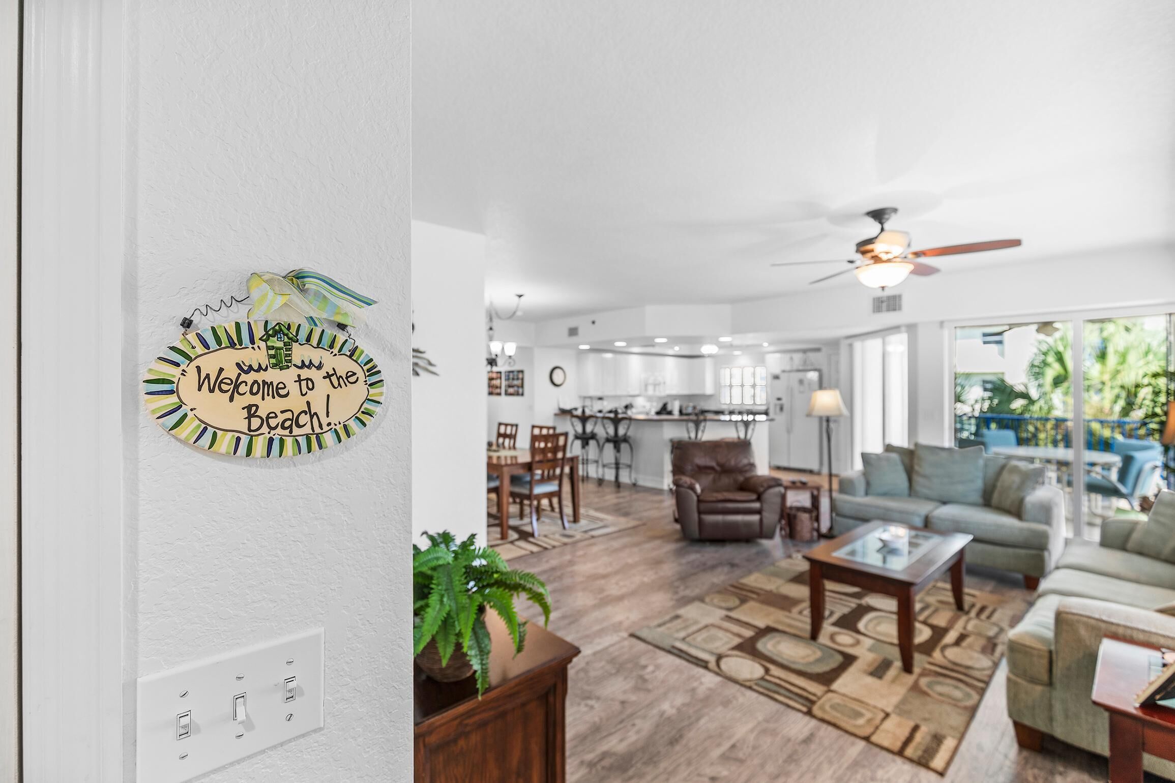 Ein geräumiger Wohnbereich mit einer Küche im Hintergrund, einem Deckenventilator und einem Schild an der Wand mit der Aufschrift 'Welcome to the Beach!'.