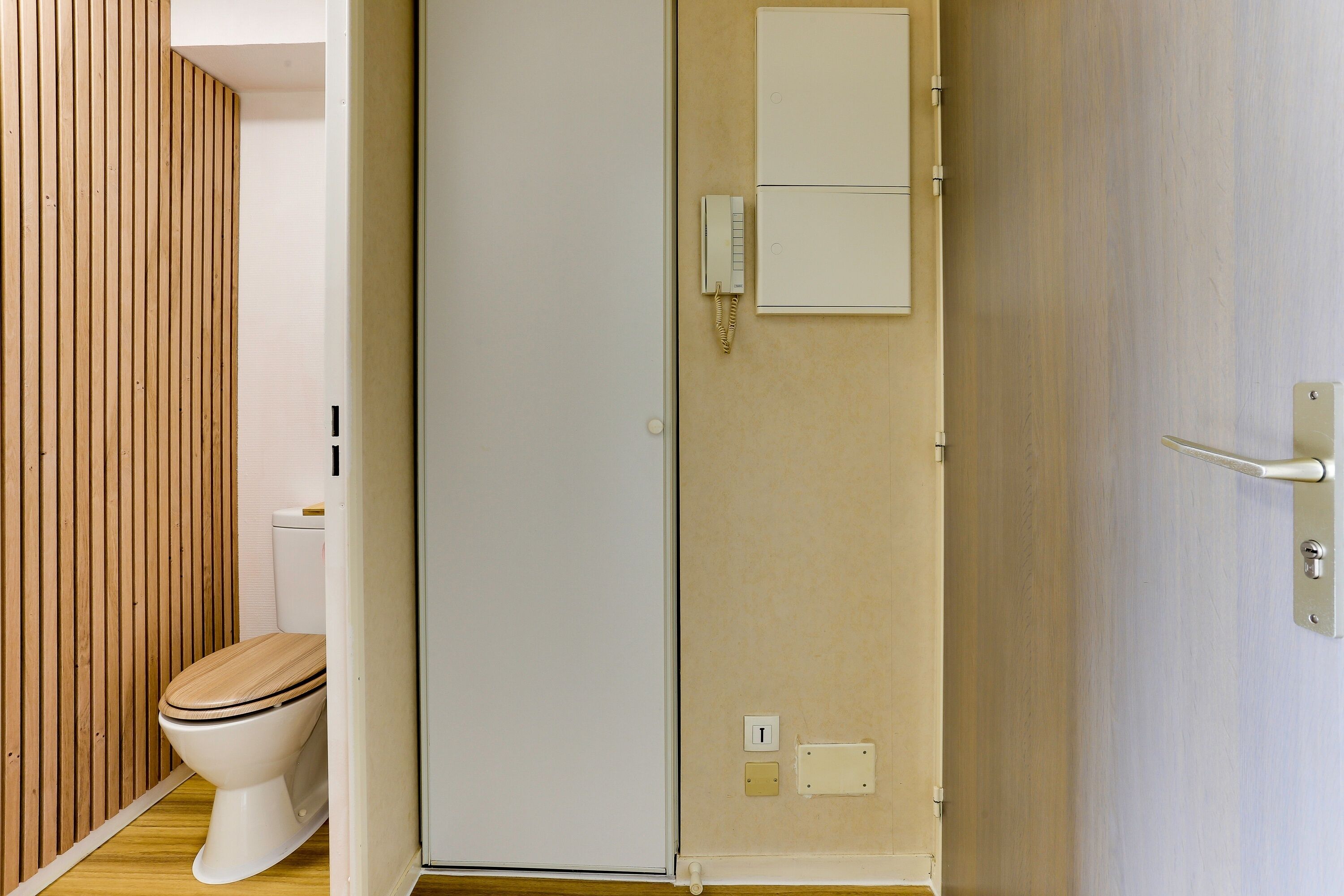 Une salle de bain avec des toilettes, un mur recouvert de panneaux en bois et une porte blanche dotée d’un interrupteur et de prises électriques.