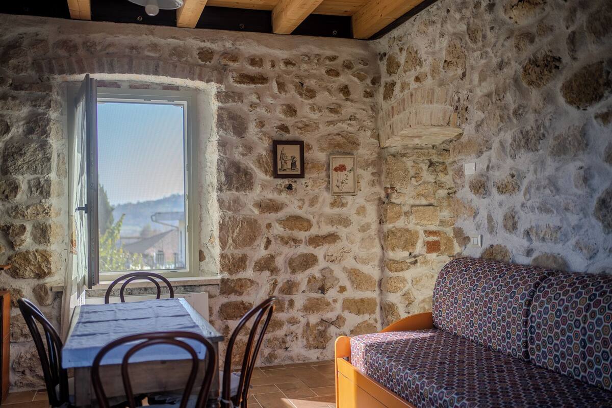 A stone-walled room with a dining table and chairs, a patterned sofa, and a window with a view.