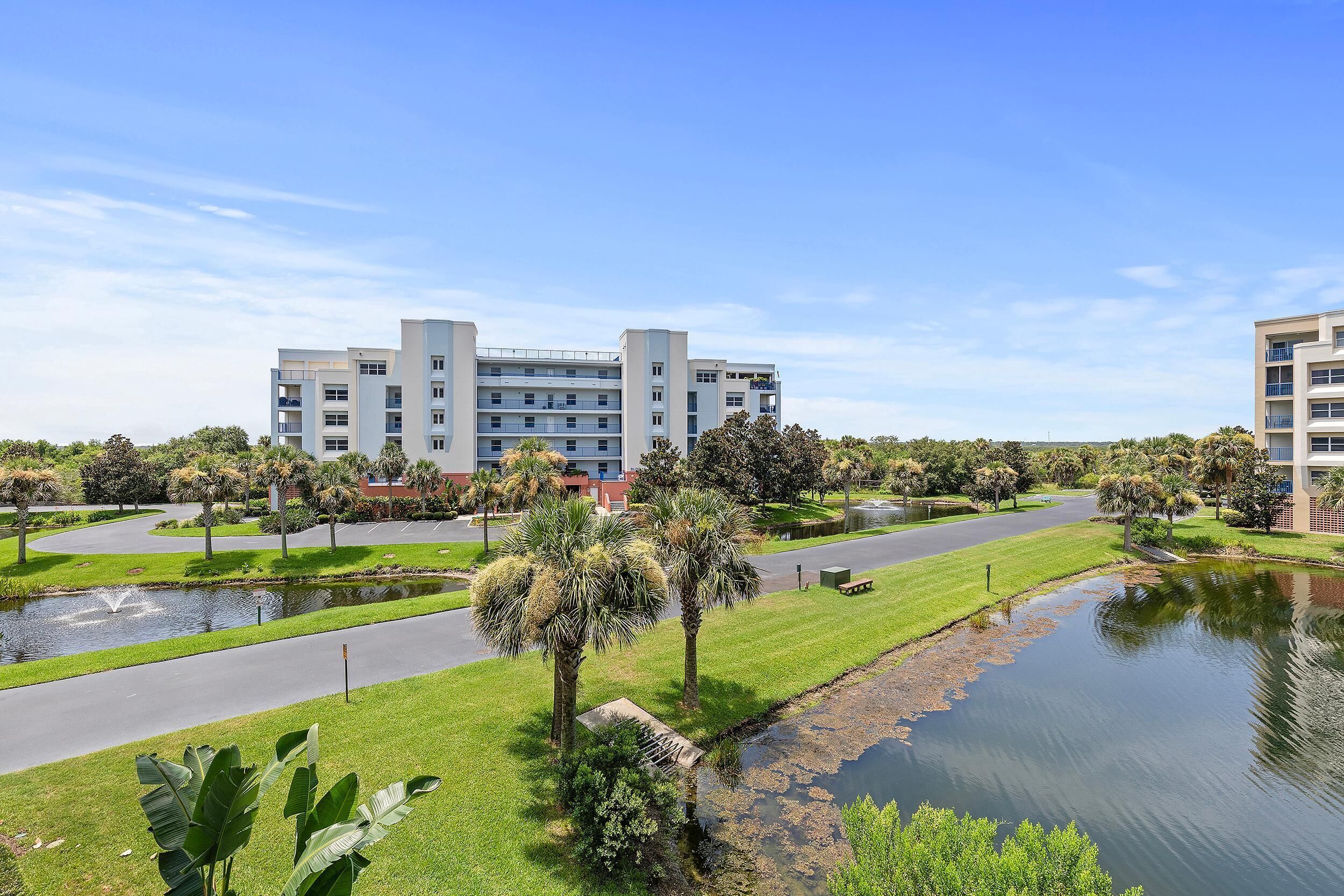 A modern building with multiple floors, surrounded by a well-maintained park with palm trees and a pond.