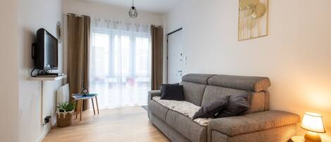 Un salon moderne avec un canapé gris, une petite table, un téléviseur posé sur une étagère et une lampe sur une table d’appoint.