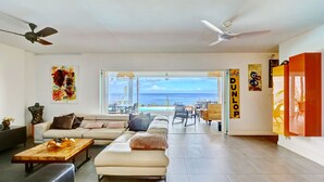 A spacious living room with a large sofa, a coffee table, and a view of the ocean. There are ceiling fans, artwork on the walls, and a sign that reads "DUNLOP".