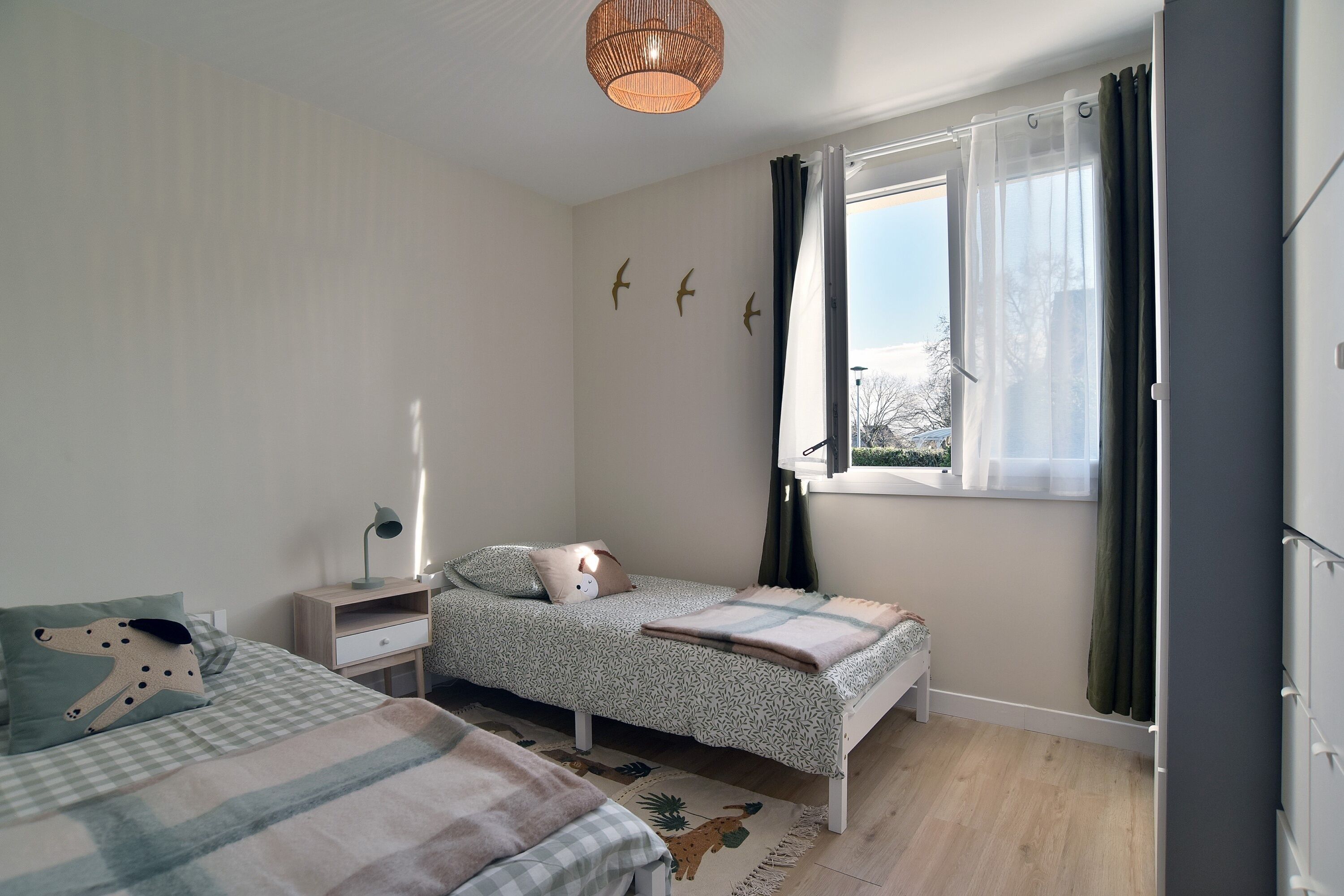 A bedroom with two beds, a window with curtains, a bedside table with a lamp, and wall art.
