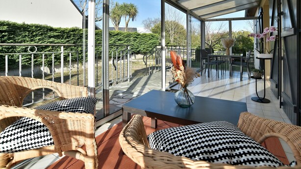A covered outdoor seating area with wicker furniture, a table with a vase, and a view of the garden.