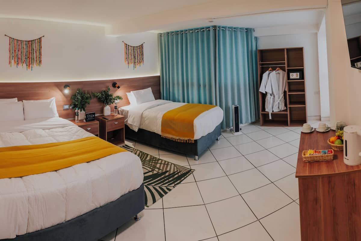 A hotel room with two beds, a wooden headboard, a bedside table with a lamp, a mirror, a wardrobe, and a small kitchenette area.