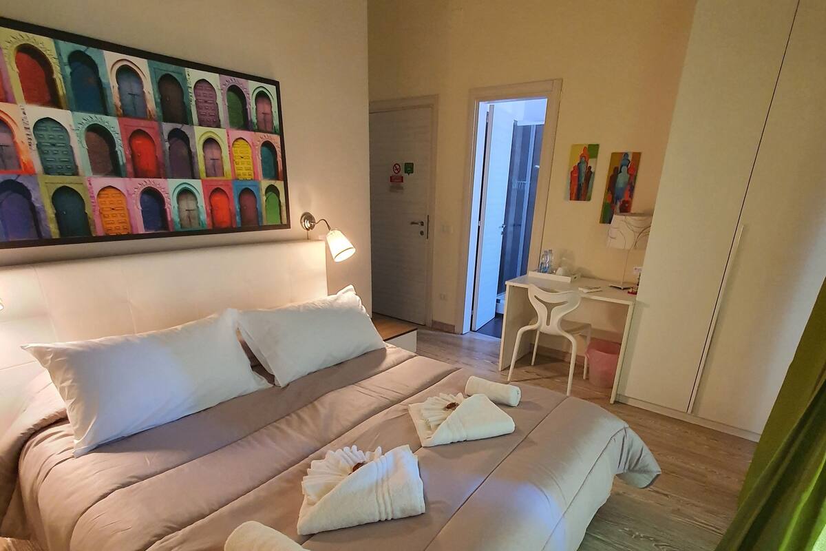 A hotel room with a large bed, a desk, and a chair. There is a painting on the wall and a door with a sign.