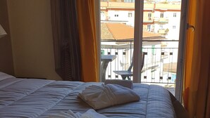 Habitación de hotel con cama, dos toallas blancas y vista a edificios a través del ventanal.