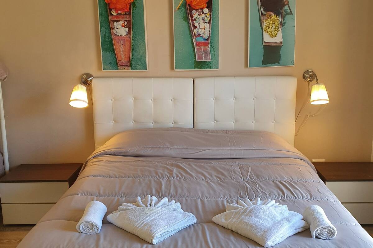 A hotel room with a large bed, two wall-mounted lamps, and three paintings depicting boats.