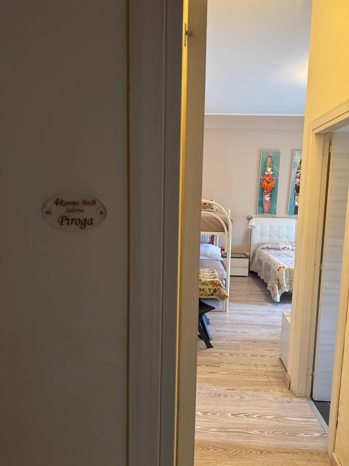 A hallway leading to a bedroom with a bed, a nightstand, and a wall-mounted lamp. A sign on the wall indicates the room number and location.