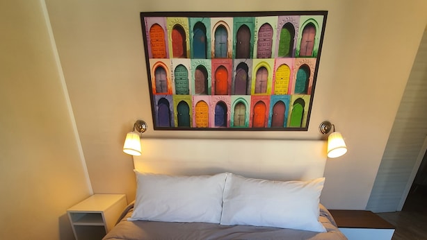 Una cama bien hecha con ropa blanca y toallas, una obra de arte llamativa en la puerta sobre ella, y dos lámparas de noche.