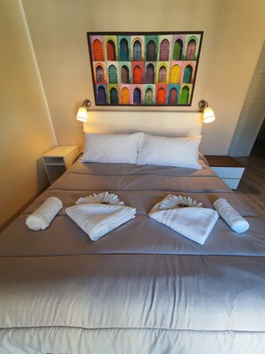 Een netjes opgemaakt bed met witte lakens en handdoeken, met bovenop een kleurrijke deurdecoratie en twee bedlampjes.