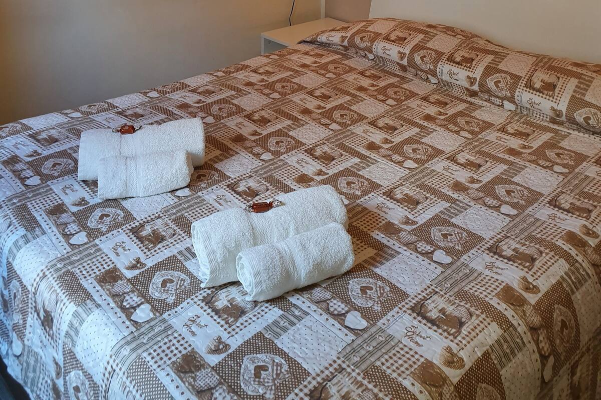 A neatly made bed with a patterned bedspread and two folded towels. There are wall-mounted artworks depicting various objects.
