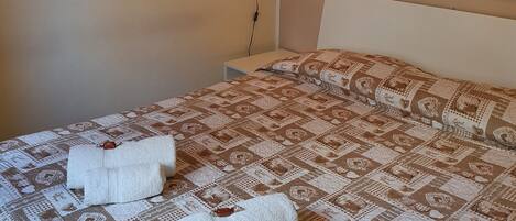 A neatly made bed with a patterned bedspread and two folded towels. There are wall-mounted artworks depicting various objects.