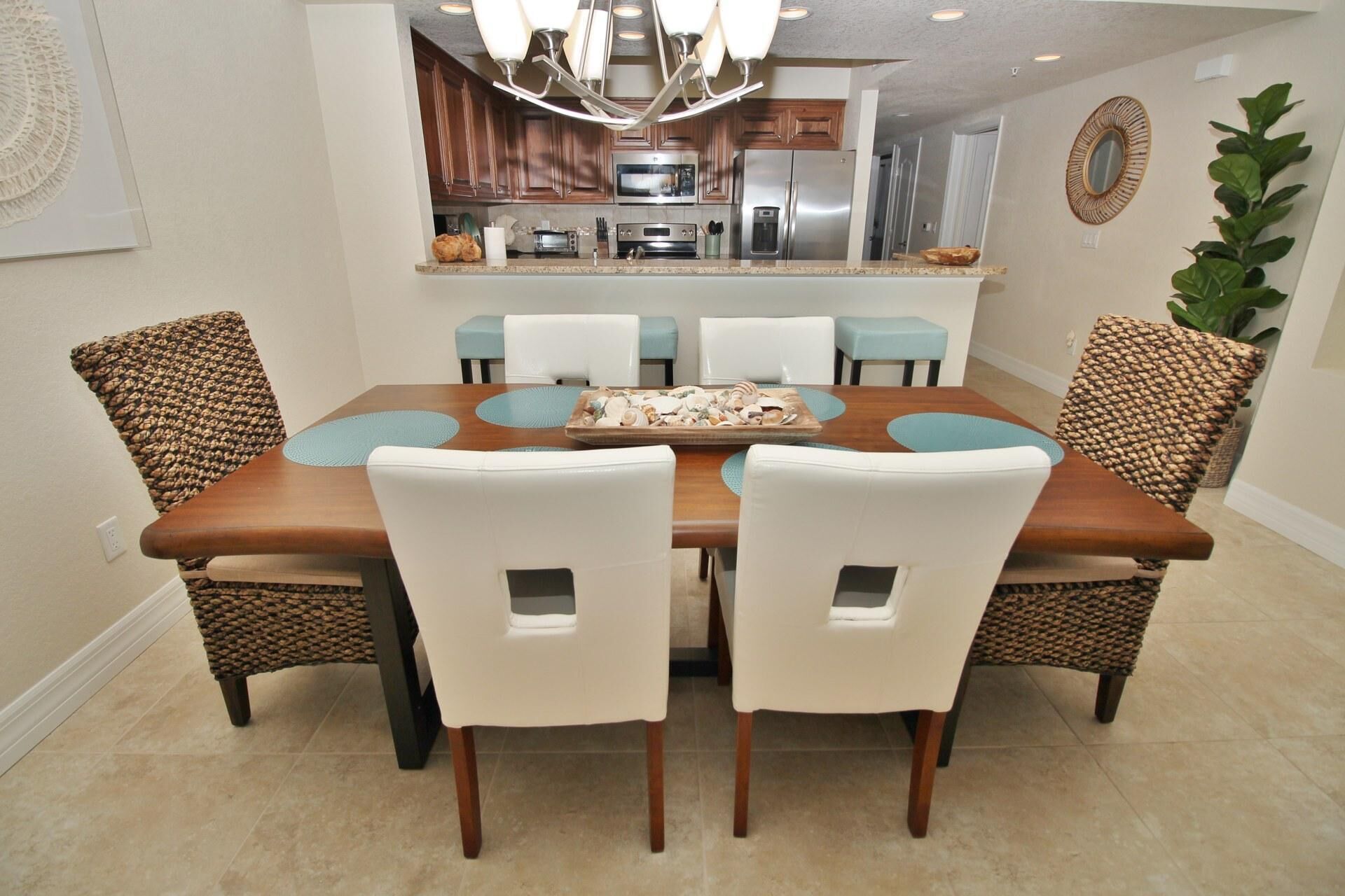 A dining area with a wooden table and white chairs, a kitchen in the background, and a potted plant on the right.