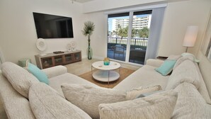 Ein Wohnzimmer mit einem großzügigen Ecksofa, einem Flachbildfernseher, einem kleinen runden Couchtisch und Blick auf einen Balkon mit Gebäuden im Hintergrund.