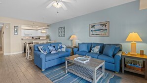 Un salon moderne avec un canapé bleu, une table basse et un coin repas, la cuisine étant visible au fond.