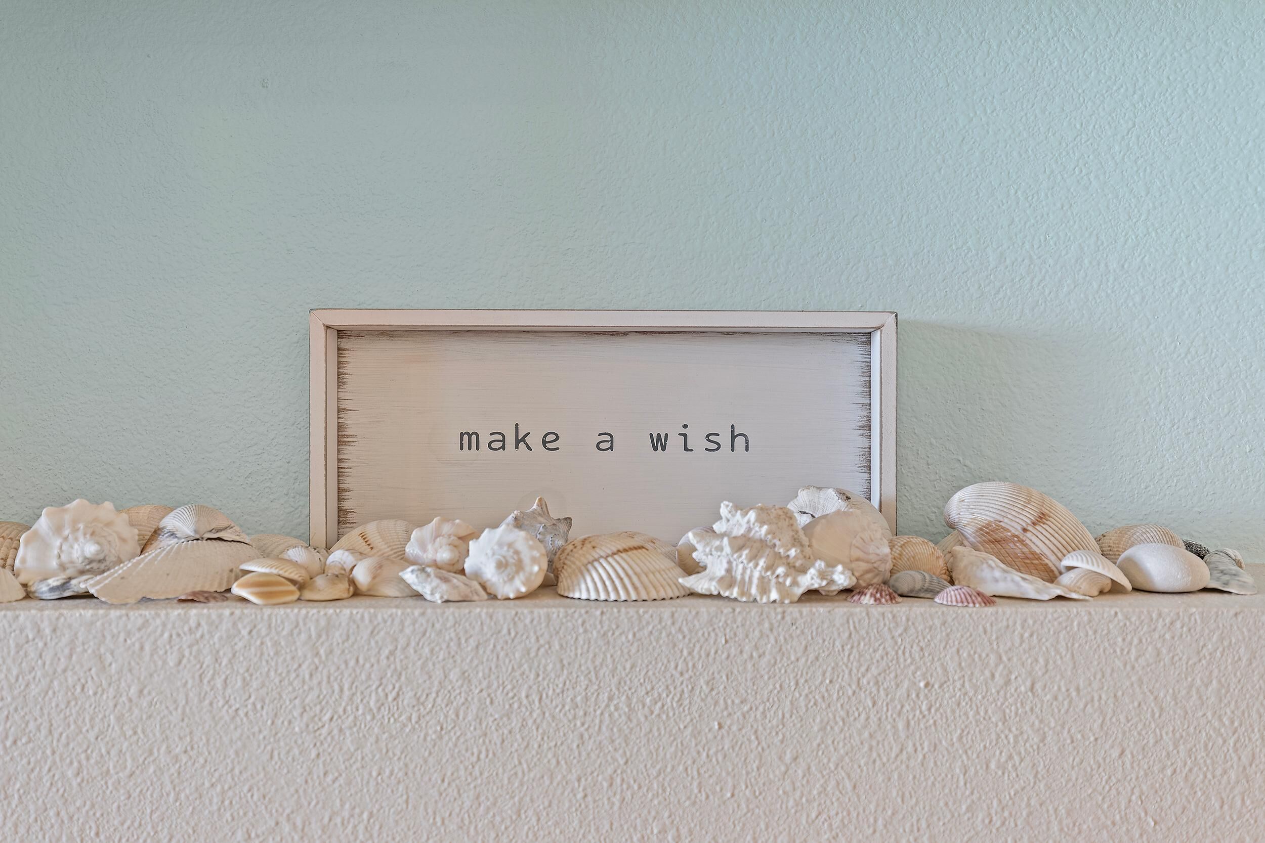 Un cartel en marco con el texto 'make a wish' rodeado de conchas marinas sobre una pared con textura.