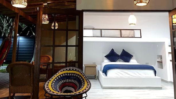 Un dormitorio moderno con una cama grande, una mesita de noche pequeña y una ventana. Hay una silla de mimbre y una lámpara colgante sobre la cama.