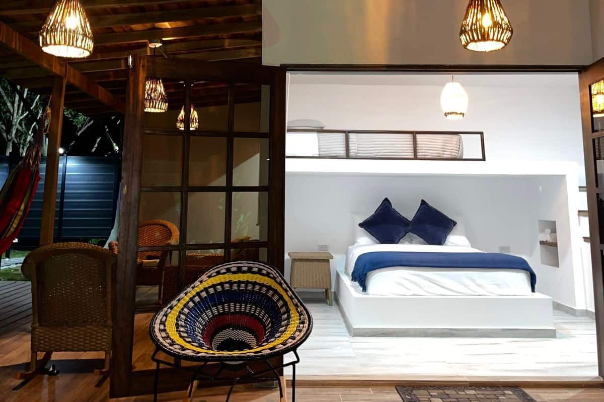 Un dormitorio moderno con una cama grande, una mesita de noche pequeña y una ventana. Hay una silla de mimbre y una lámpara colgante sobre la cama.
