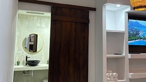 Un baño moderno con un espejo grande, un lavabo negro y una puerta de madera. Se observa una estantería blanca con un televisor que muestra una vista panorámica.