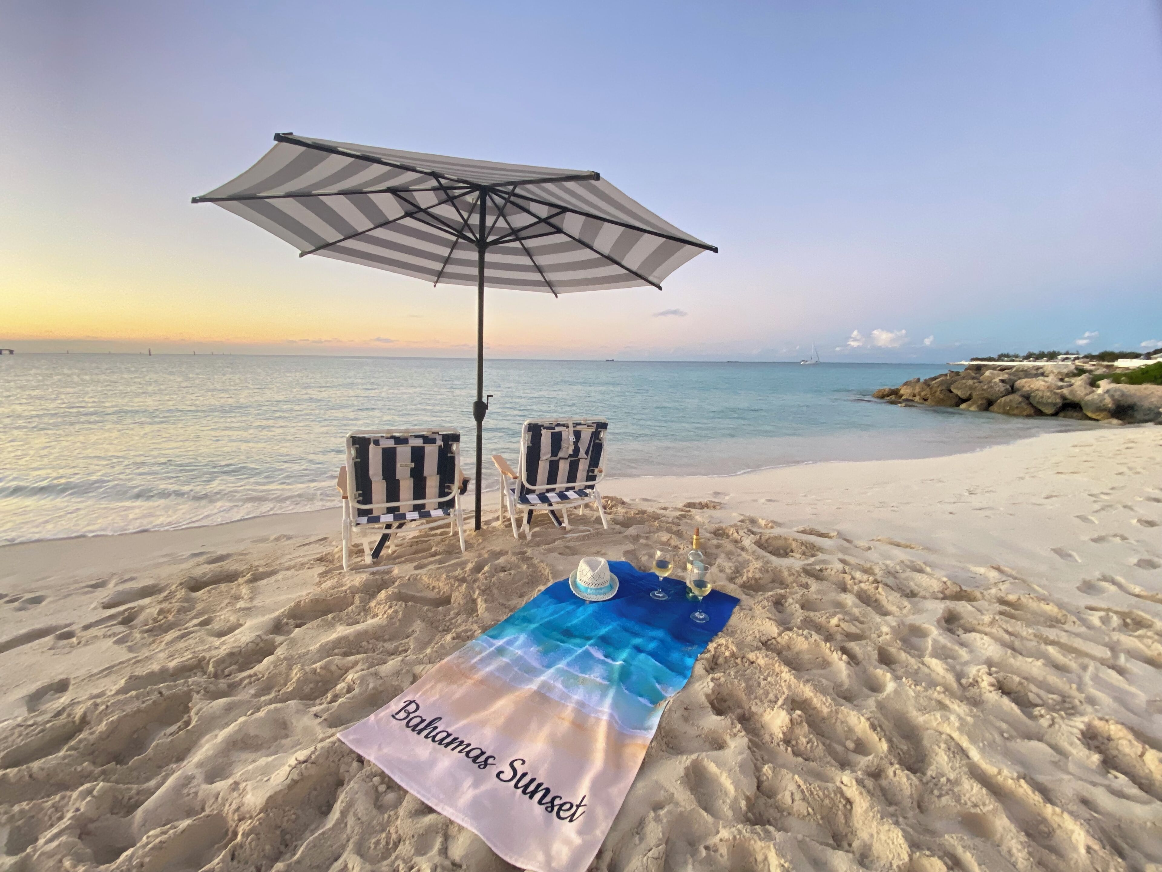 Une scène de plage avec deux chaises de plage à rayures, un parasol et une serviette sur laquelle est inscrit le texte « Bahamas Sunset ».
