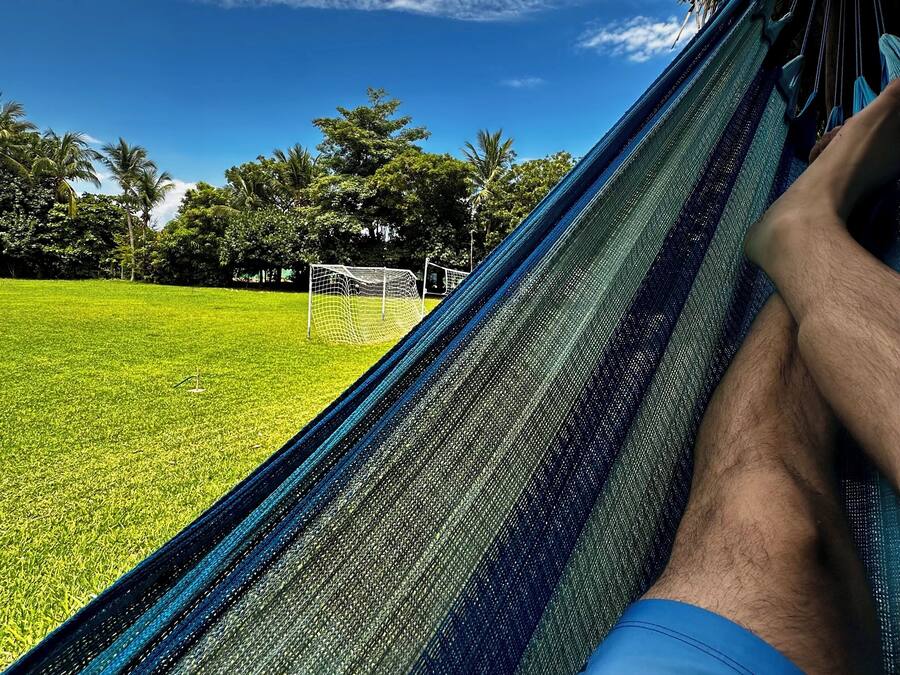 Uma rede com uma pessoa deitada, com vista para um campo gramado e árvores sob um céu parcialmente nublado.