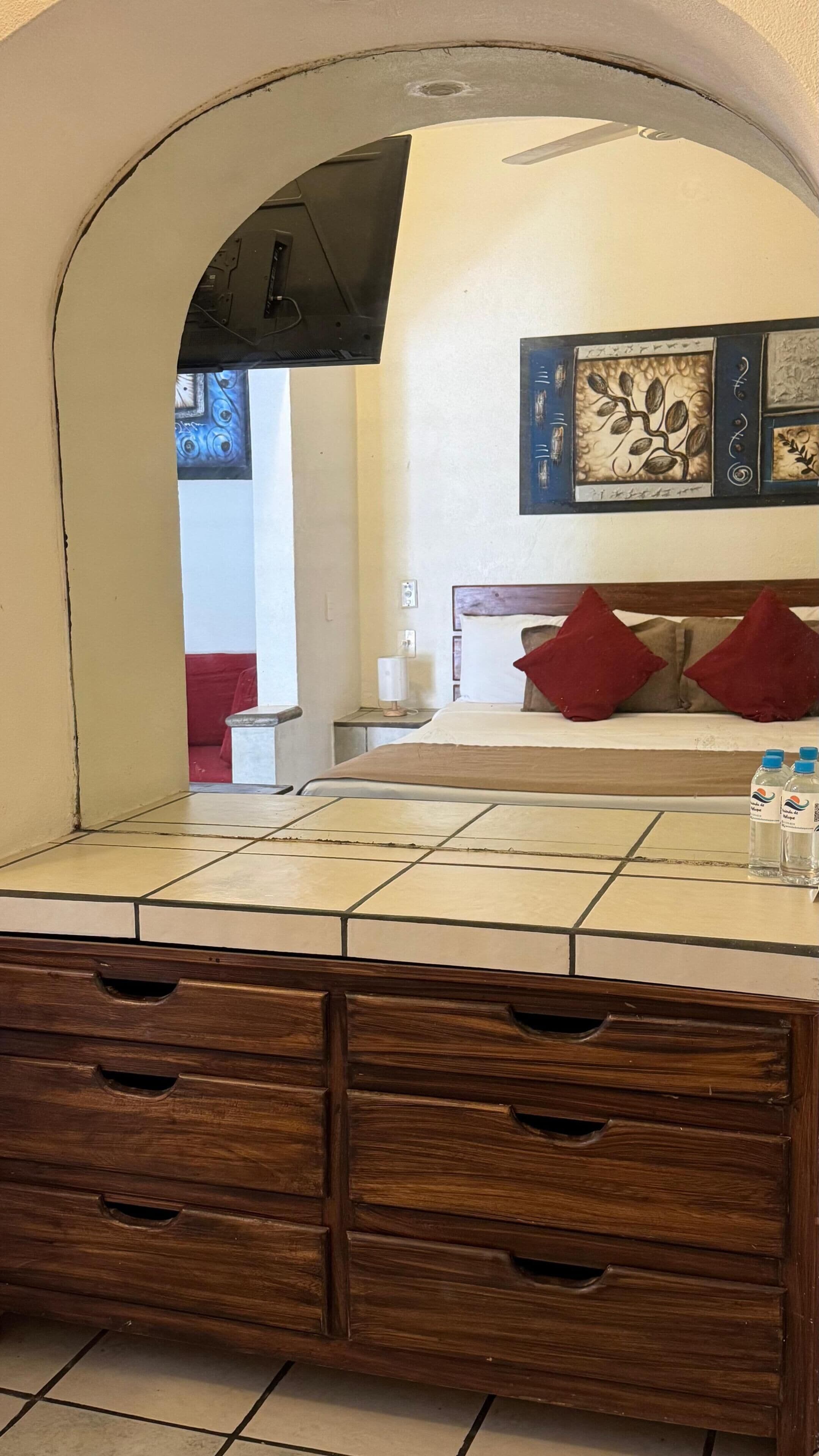 A bathroom with a tiled vanity, wooden drawers, a mirror, and a bed with red pillows in the background.