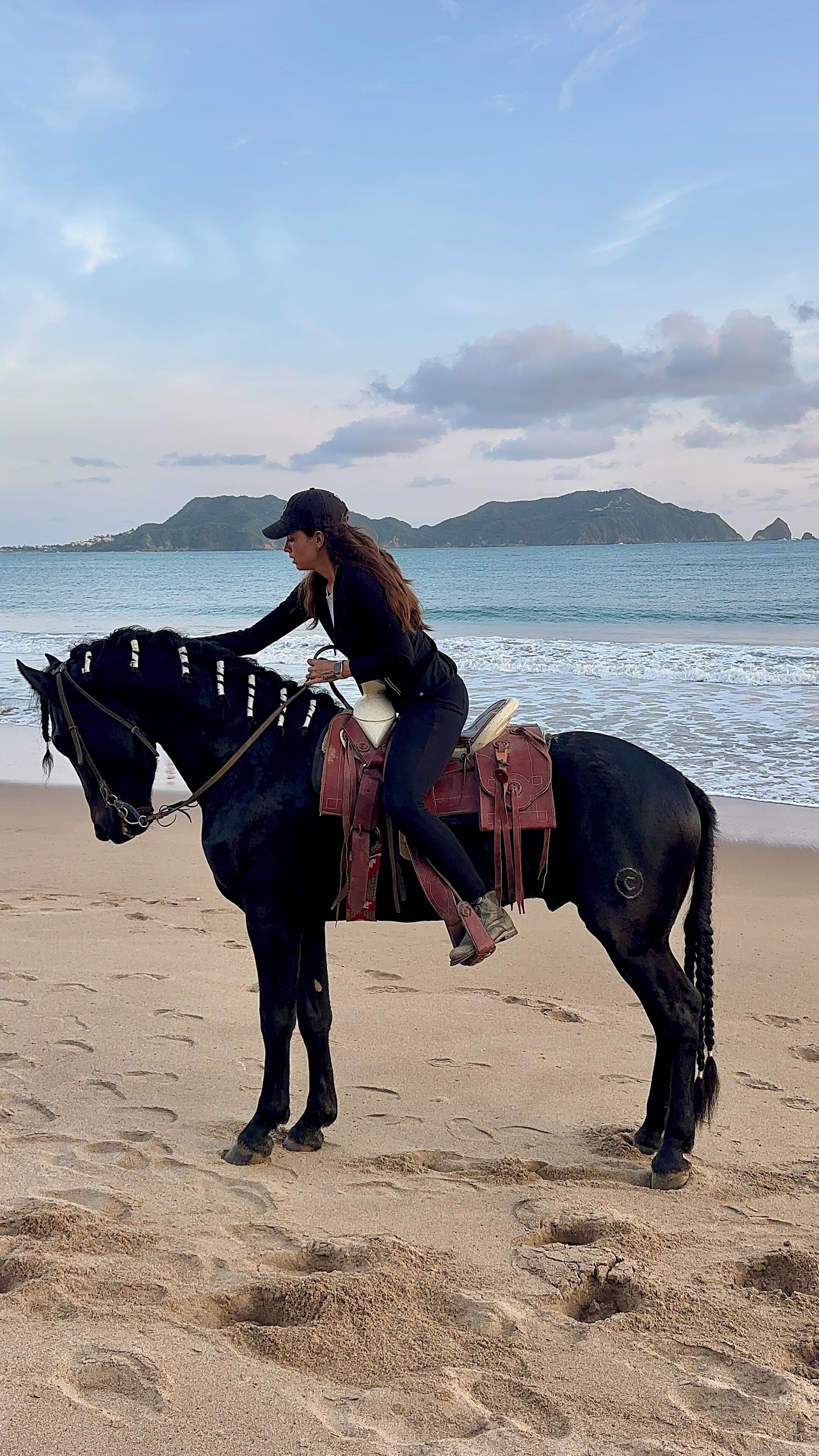 Une personne à cheval sur une plage, avec une étendue d'eau et des collines en arrière-plan.