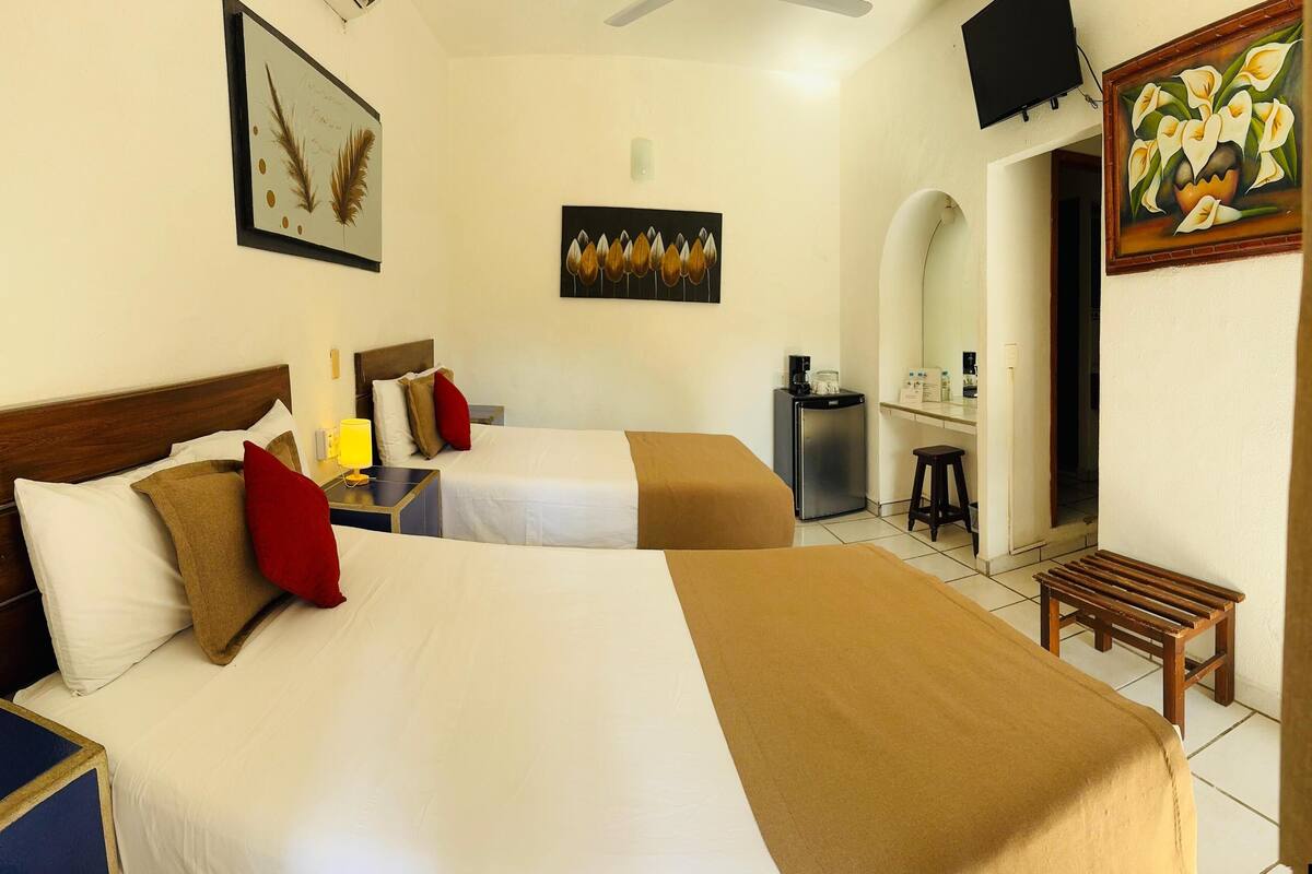 Una habitación de hotel con dos camas, un escritorio, una silla, un televisor y obras de arte en las paredes.