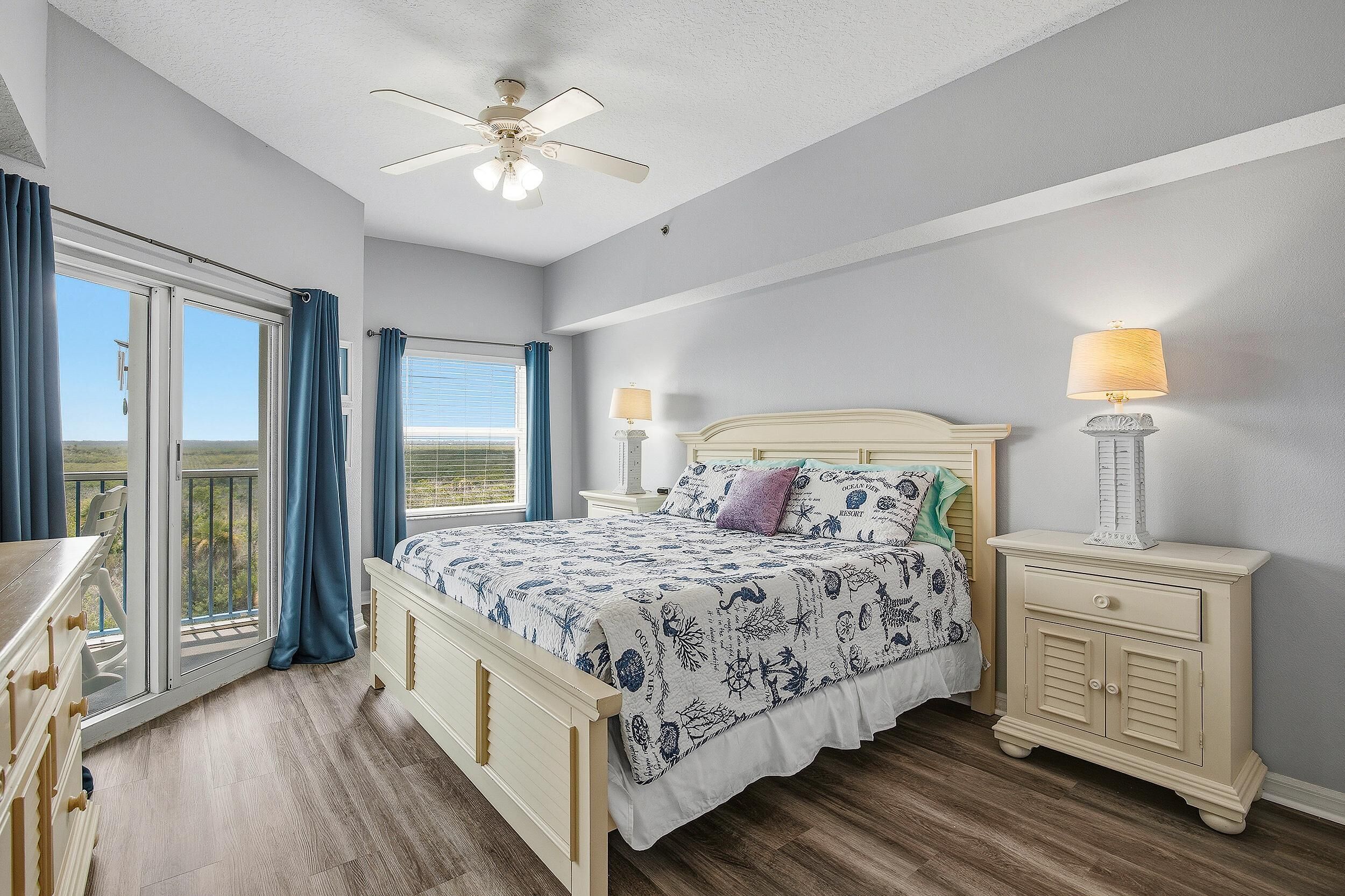 A bedroom with a bed, two bedside tables, a ceiling fan, and a view through a large window.