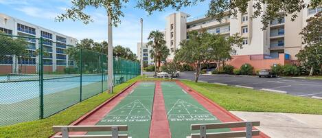 A shuffleboard court with a green surface and red borders, benches on either side, and a chain-link fence enclosing a tennis court.