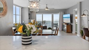 A modern living room with a dining area, large windows, a chandelier, and a television. - Ponce de Leon Towers 401 (New Smyrna Beach)