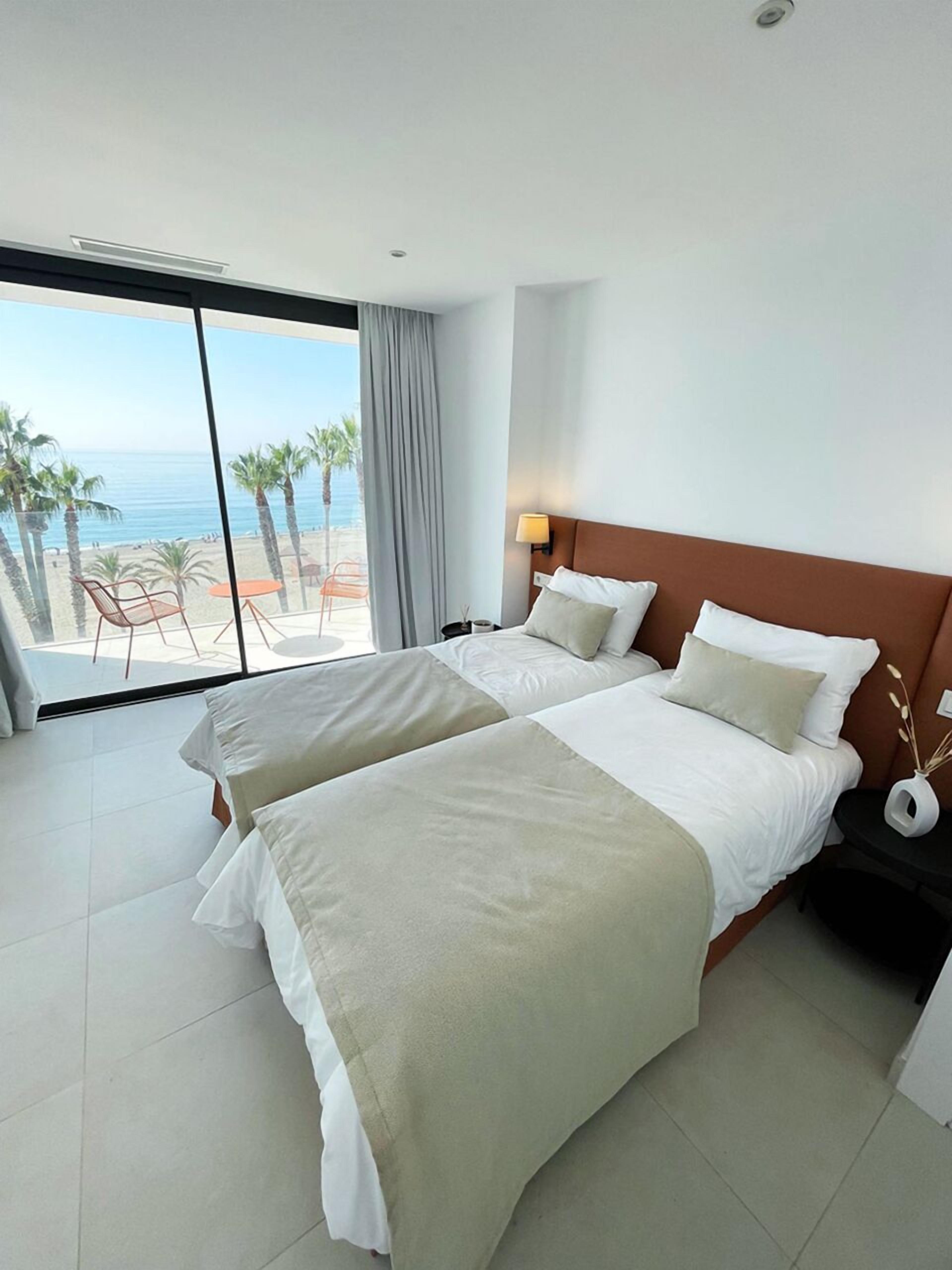 Ein Hotelzimmer mit zwei Betten, einem großen Fenster mit Blick auf den Strand und Palmen sowie einem kleinen Tisch mit einer Lampe.