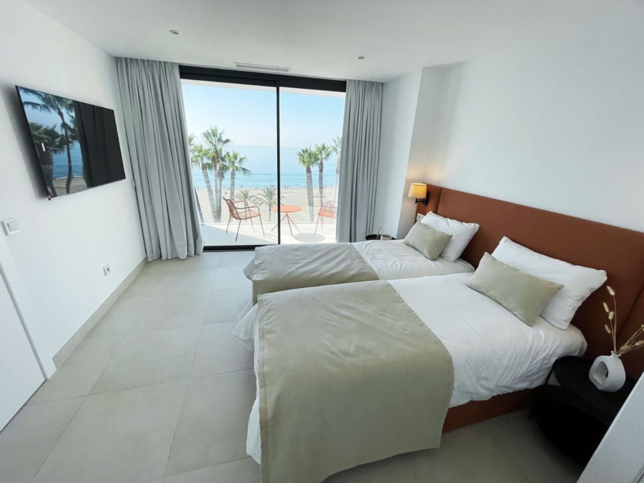 A hotel room with two beds, a TV, a balcony with a view of the beach, and a small table with chairs.