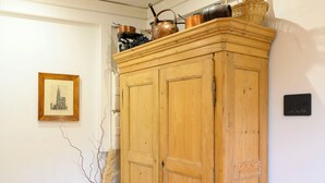 Ein Holzkabinett mit Kupferkochgeschirr und einem Weidenkorb auf der Oberseite, ein gerahmtes Bild an der Wand und eine grüne Vase mit getrockneten Ästen.