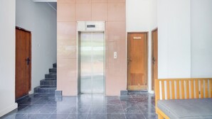 A hallway with a central elevator, a wooden door to the left, and a bench to the right. - Hotel O Palmerah Near Binus University Formerly Wisma Bango (Jakarta)