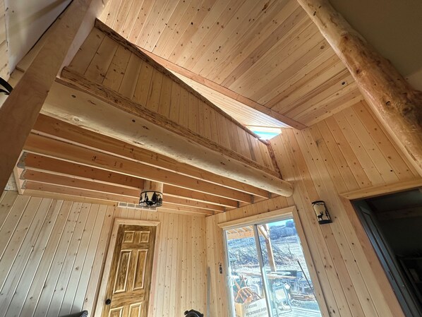 A wooden cabin interior with a sloped ceiling, a door, and a window with a view of an outdoor area.