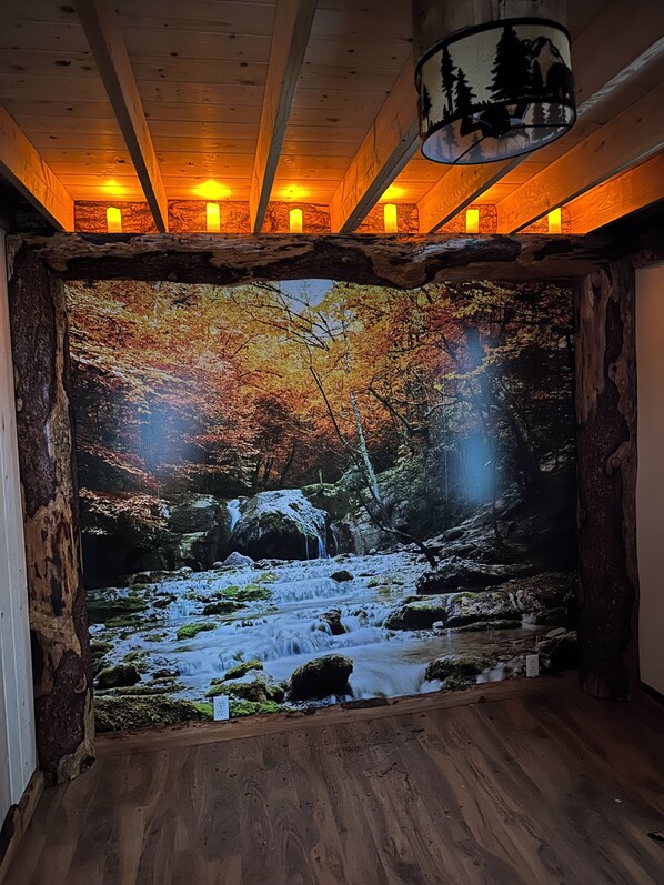 Habitación con un gran mural en la pared de un arroyo en el bosque, vigas de madera en el techo y una lámpara colgante.