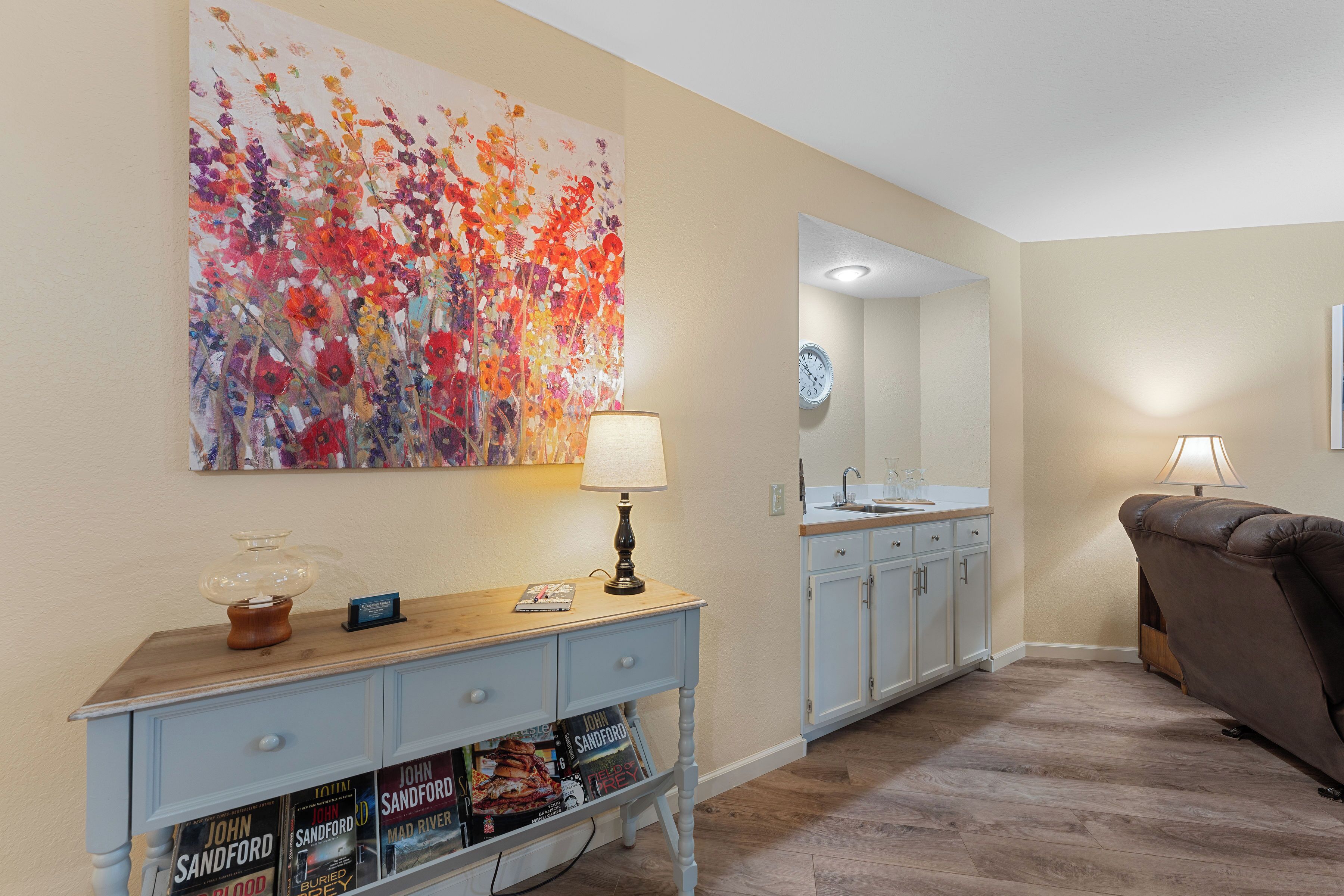 A living room with a light blue console table, a large floral painting, a sofa, and a kitchen area in the background.