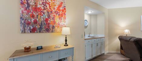A living room with a light blue console table, a large floral painting, a sofa, and a kitchen area in the background.