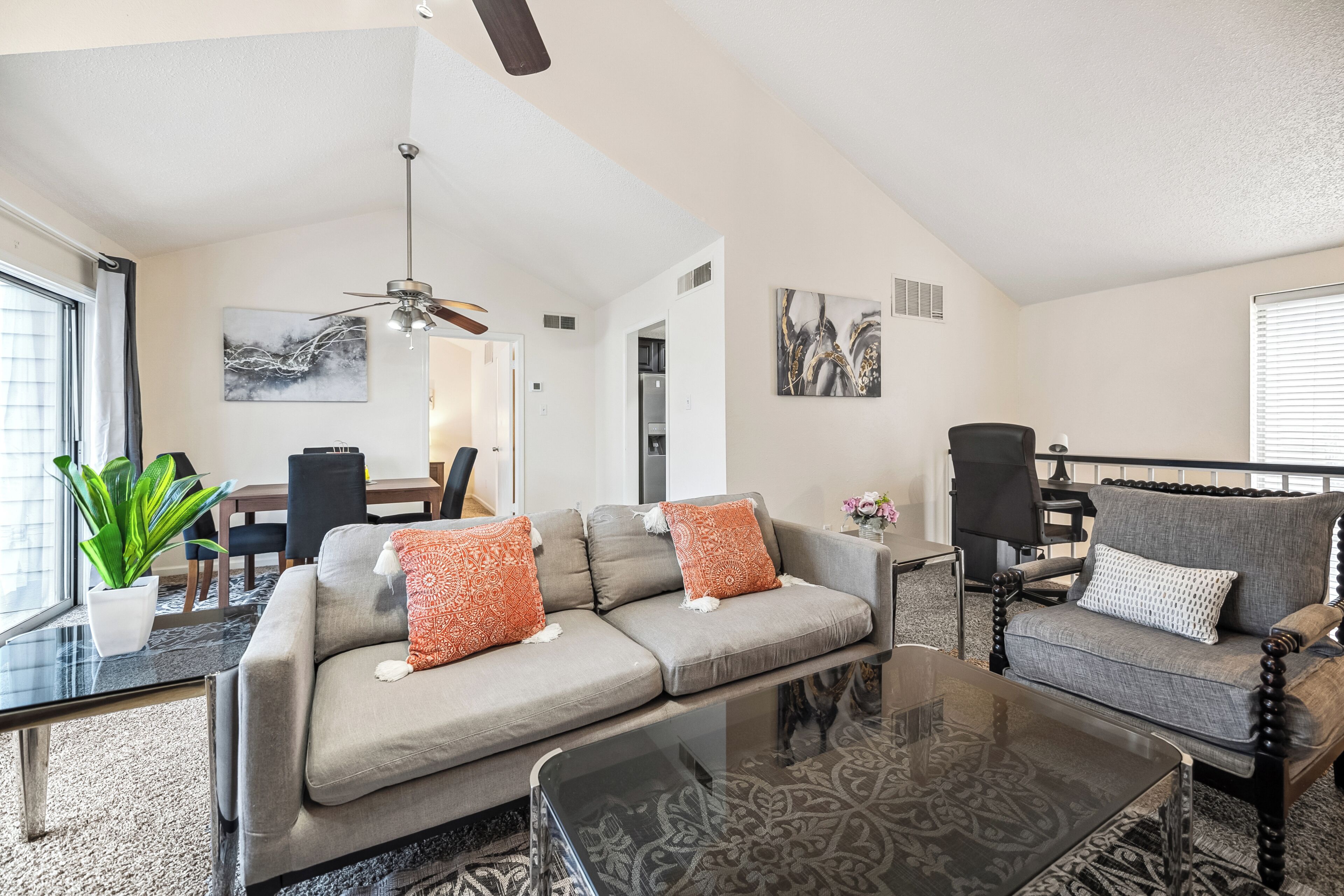A modern living room with a sofa, armchair, and coffee table. There is a dining area with a table and chairs, and a kitchen with a refrigerator.