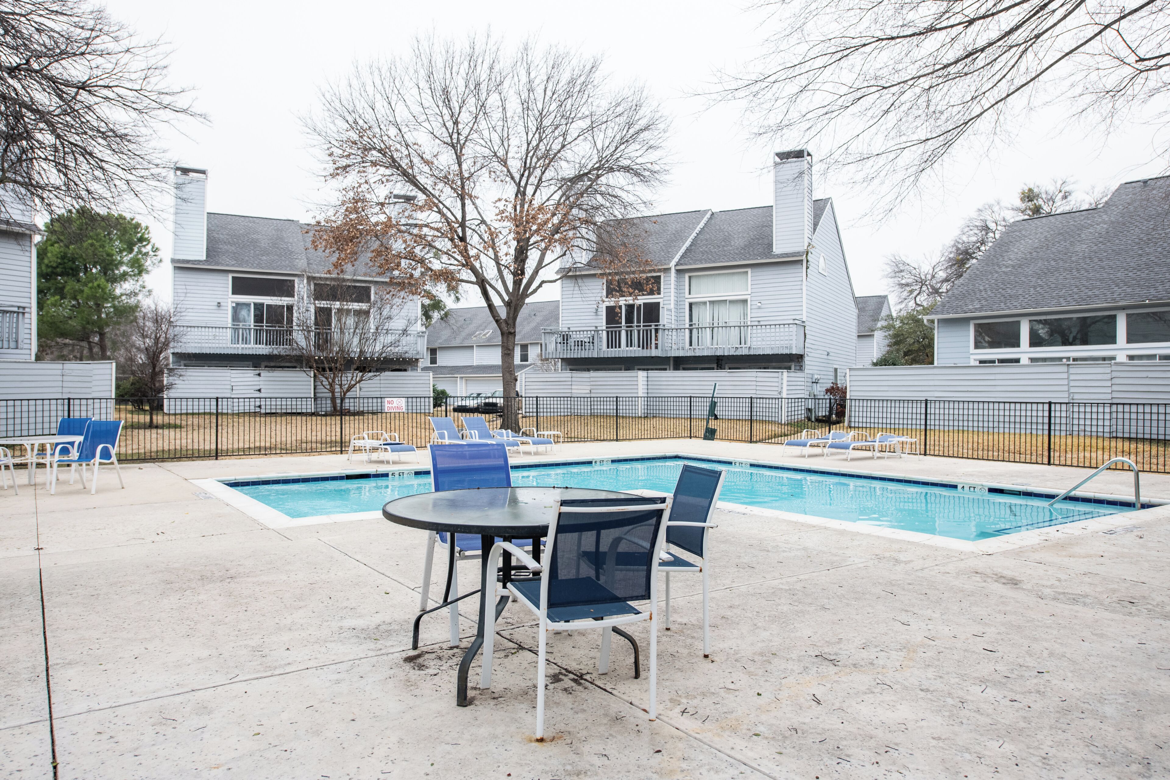 A residential swimming pool with lounge chairs and a table, surrounded by a fence and houses in the background.