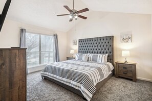 Ein Schlafzimmer mit einer gesteppten Kopfteil, einem Bett mit gestreifter Bettwäsche, einem Nachttisch mit Lampe, einem Deckenventilator und einem Fenster mit Jalousien.