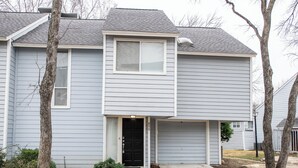 A two-story residential building with a garage, a front door, and a small front yard with a tree and shrubs.